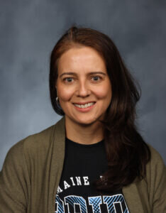 Woman with medium length brown hair wears a black t-shirt with blue letters and an olive green cardigan smiles in front of a grey background.