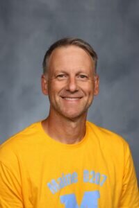 Headshot of a smiling middle-aged man with short, light brown hair, wearing a bright yellow t-shirt with Maine D207 printed in blue. He has a slight stubble and is set against a mottled gray studio background.