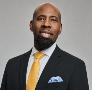 A man in a black suit, white shirt, and yellow tie smiles at the camera. He has a beard and is wearing a blue pocket square. The background is plain and gray.